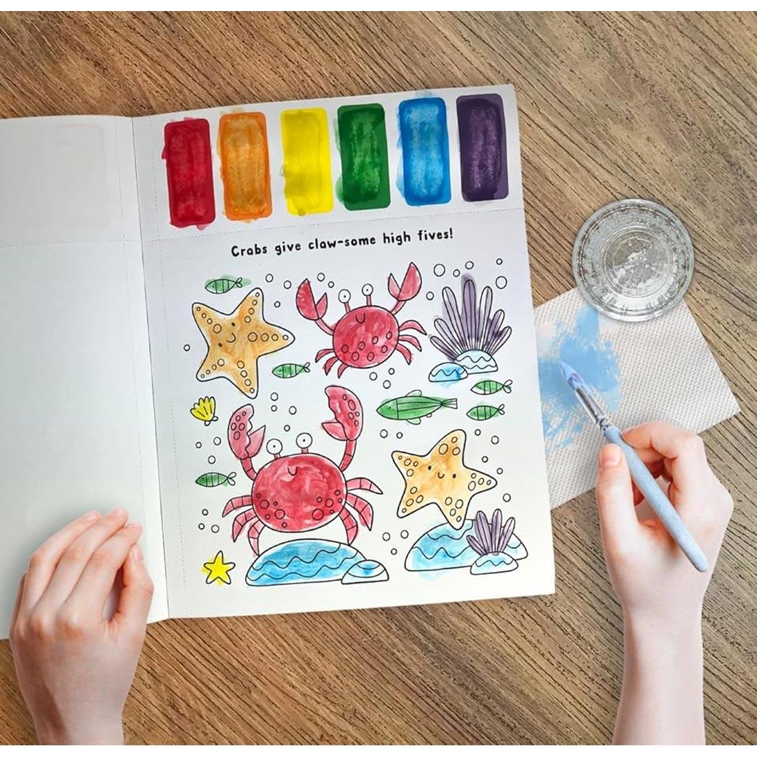 Hands holding a paintbrush with completed page from Under The Sea Paint Magic Book
