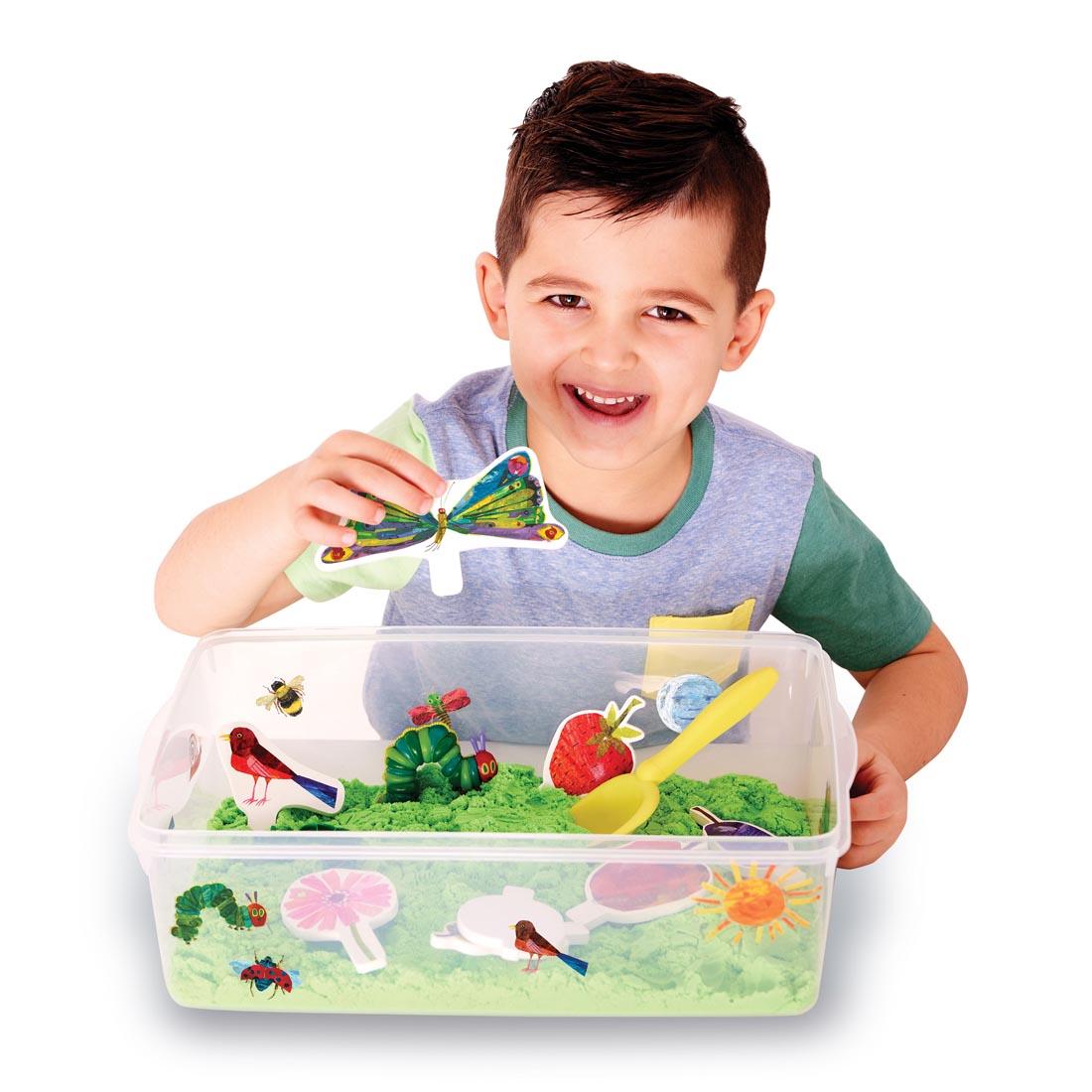 Contents of The Very Hungry Caterpillar Sensory Bin, showing an open tub with sand and various pieces, with a young boy holding a butterfly piece