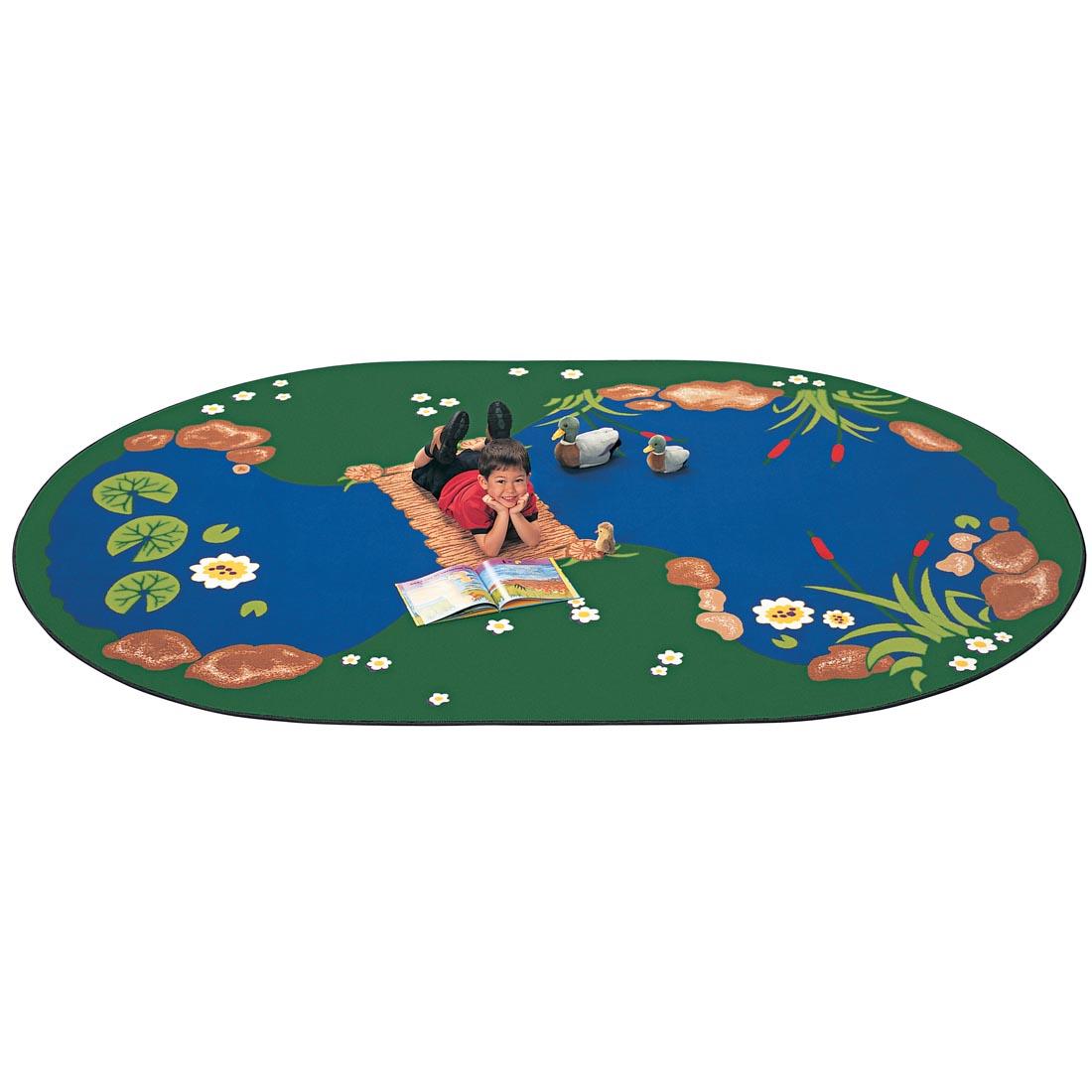 The Pond Oval Rug by Carpets For Kids with a young boy laying on the rug reading a book