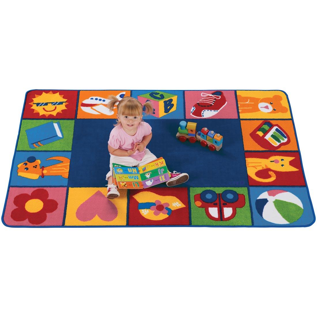 Child with a book and toy train sitting on the Toddler Blocks Rug by Carpets For Kids