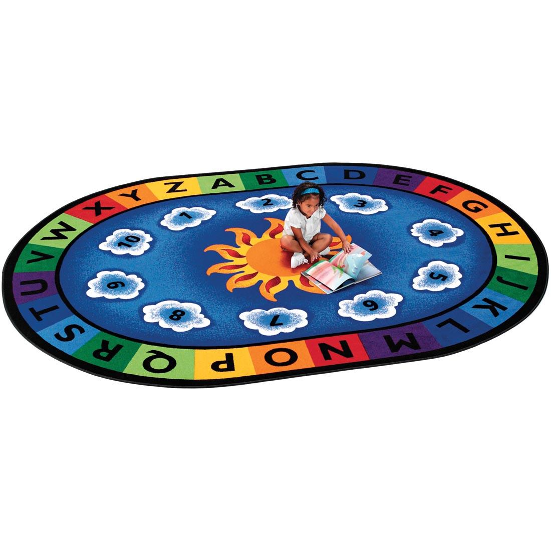 Child with a book sitting on the Sunny Day Learn & Play Oval Rug by Carpets For Kids
