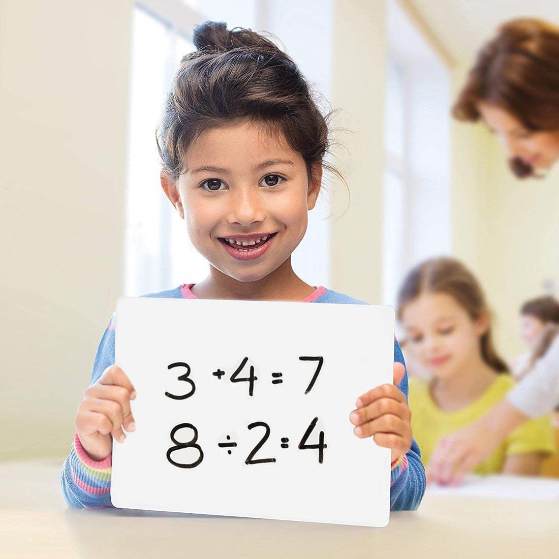 child holding a Dry Erase Lapboard with two equations written on it and a classroom scene in the background