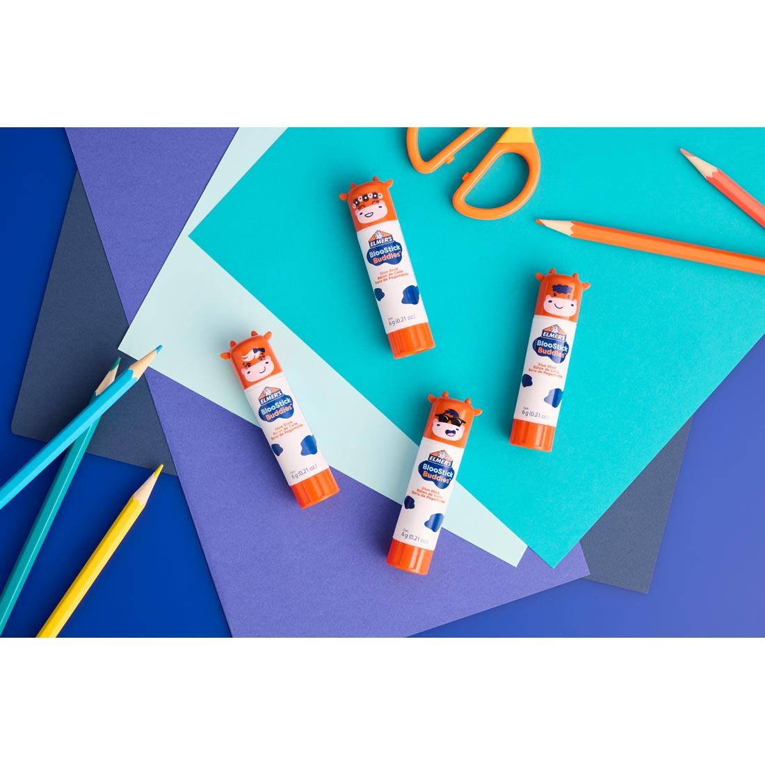 4 Elmer's BlooStick Buddies Washable Glue Sticks laying on a table with colored paper, scissors and colored pencils