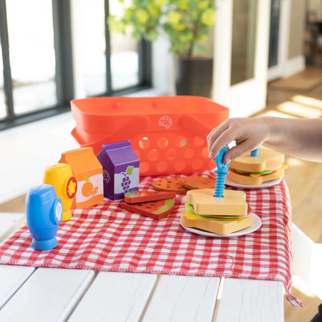 Play food, picnic basket and table cloth on a picnic table, with a hand using the "toothpick" screw to hold sandwhich together