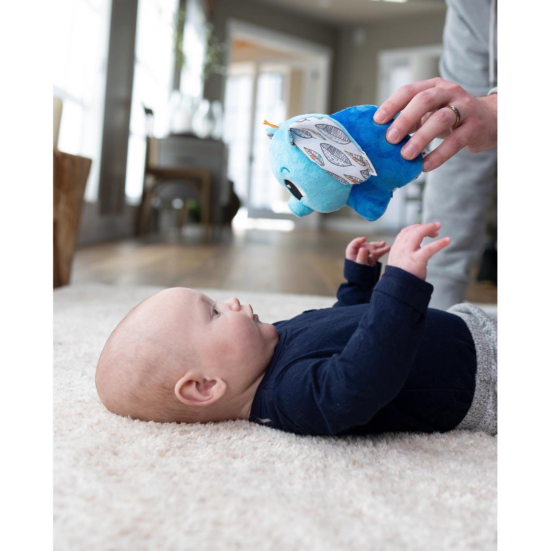 baby lying on the floor with an adult hand holding the Puffaboo Elephant By Lamaze over them