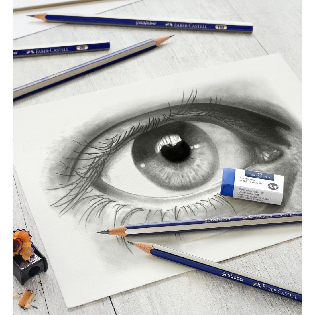 Several GoldFaber Graphite Pencils, an Eraser and a Sharpener, shown on top of a drawing of an eye