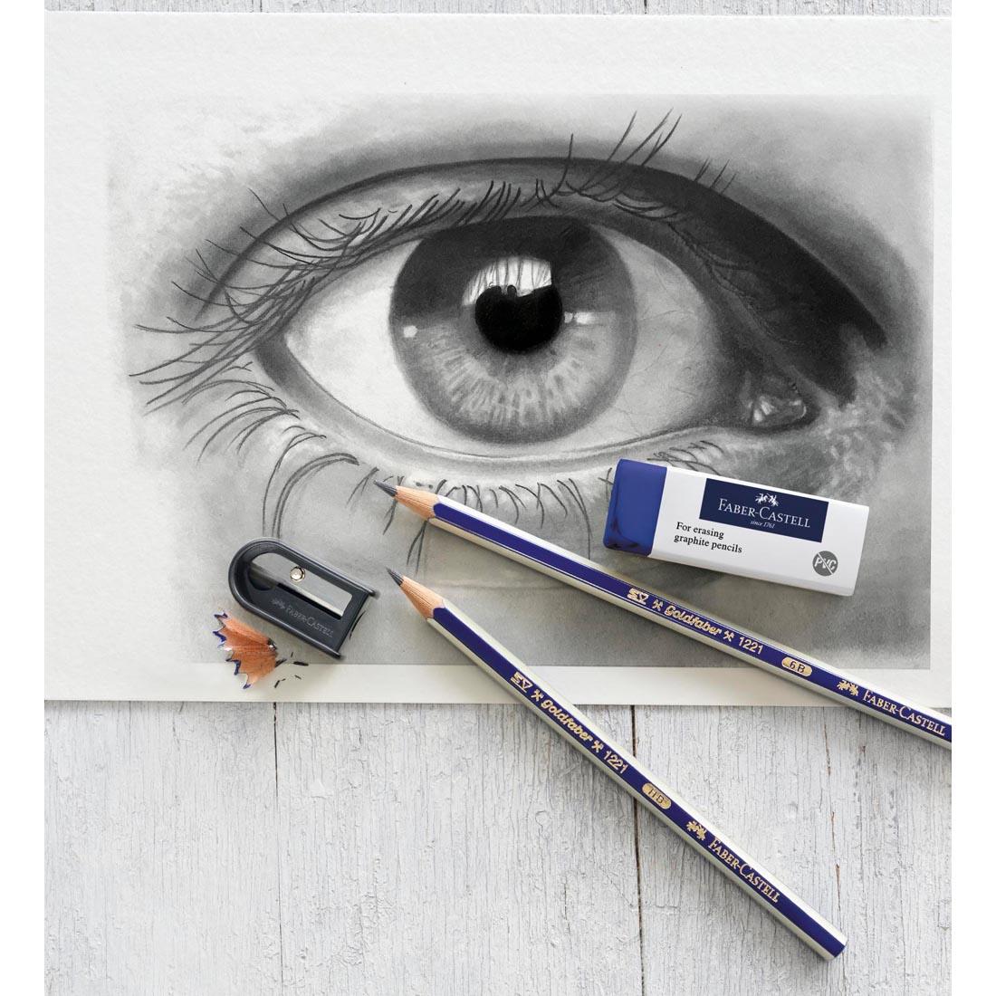 2 GoldFaber Graphite Pencils, an Eraser and a Sharpener, shown on top of a drawing of an eye
