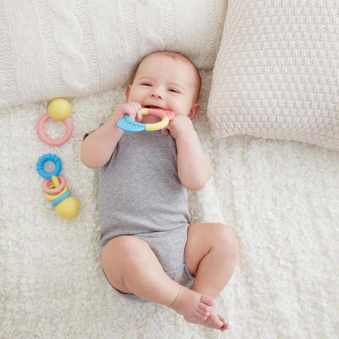 Baby lying down beside 2 of the Rattle & Teether Collection and one in their mouth