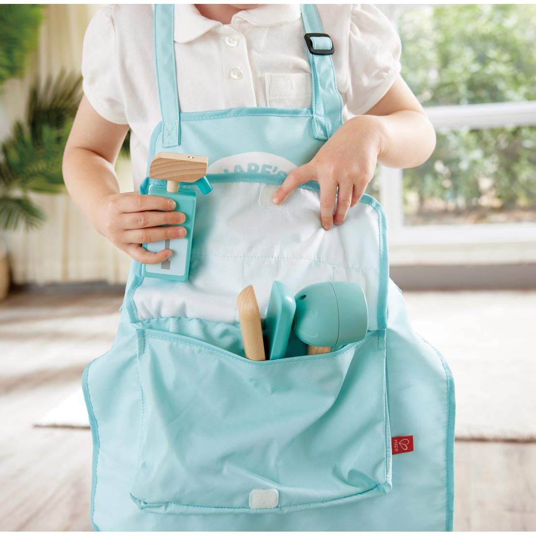 child wearing the apron holding pieces from the Super Stylish Hair Salon Set By Hape