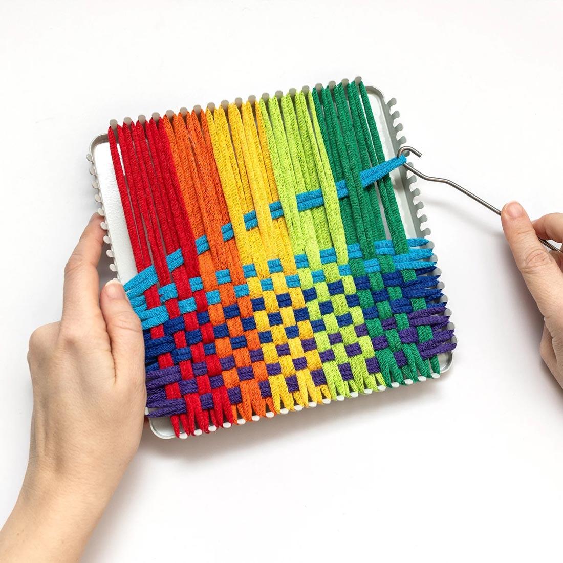 hands holding a potholder loom, and using a hook to weave an assorted color potholder