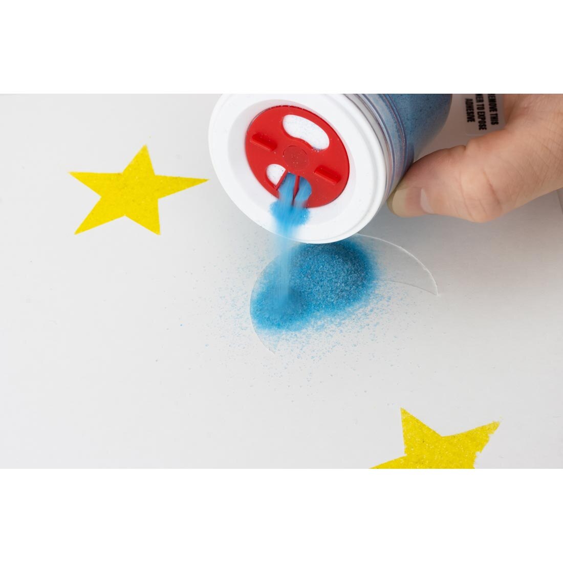 hand pouring sand onto a crescent moon shape that has been cut into a Collage Sticky Board; two stars are on either side of the moon