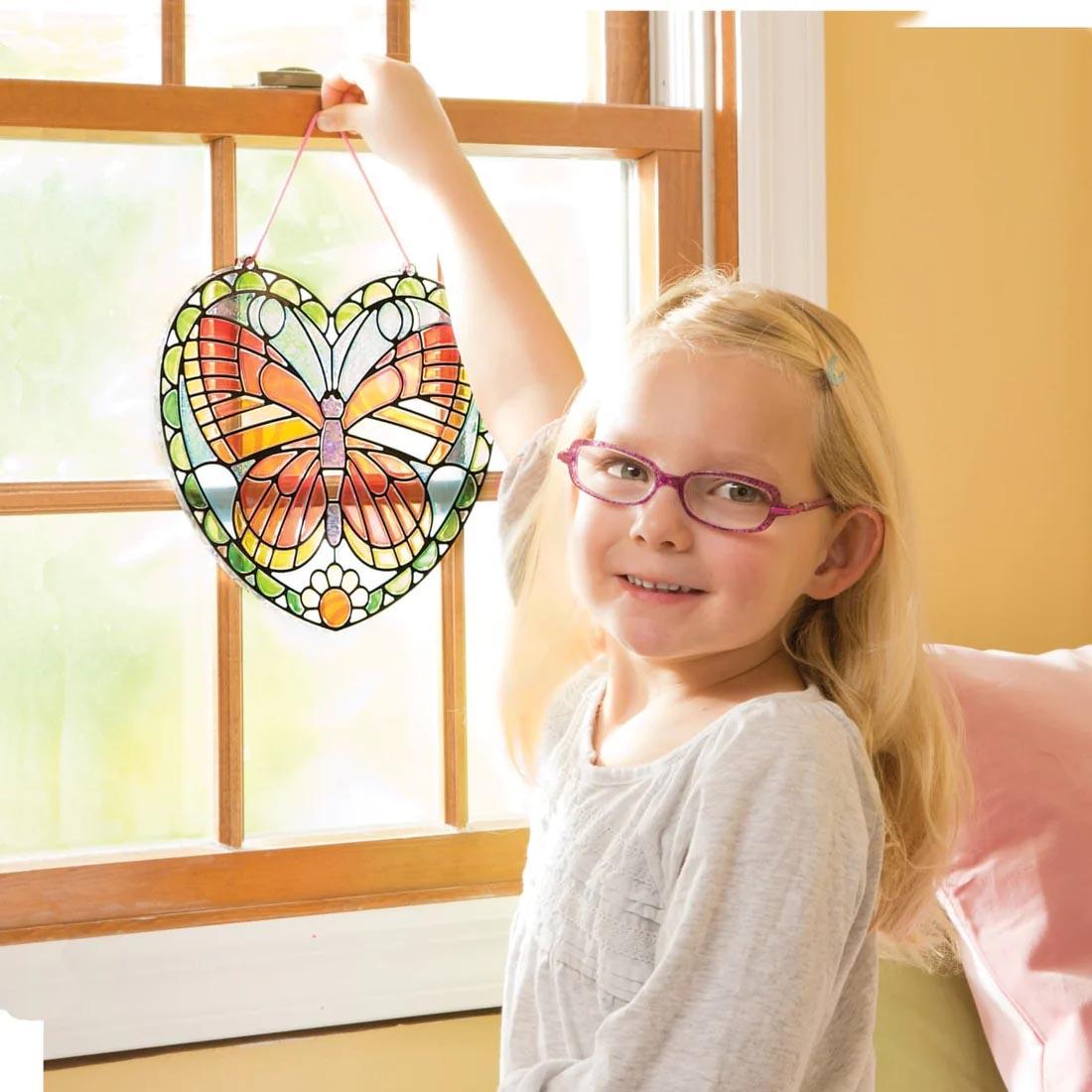 child hanging a completed Melissa & Doug Stained Glass Made Easy Butterfly in the window