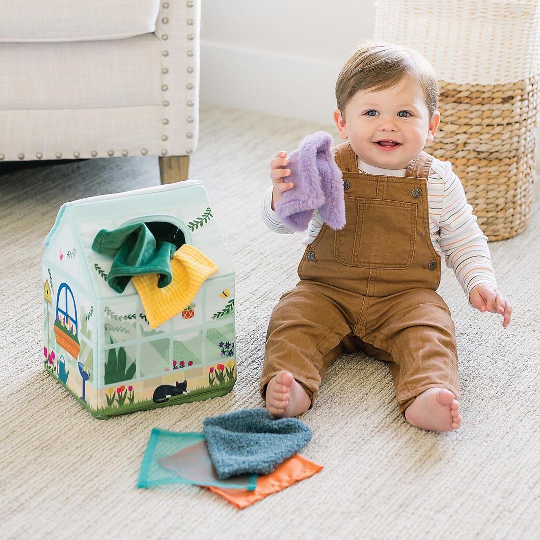 baby sitting on the floor next to the Sensory Sprouts Peek & Pull Baby Tissue Box Toy By Mindware