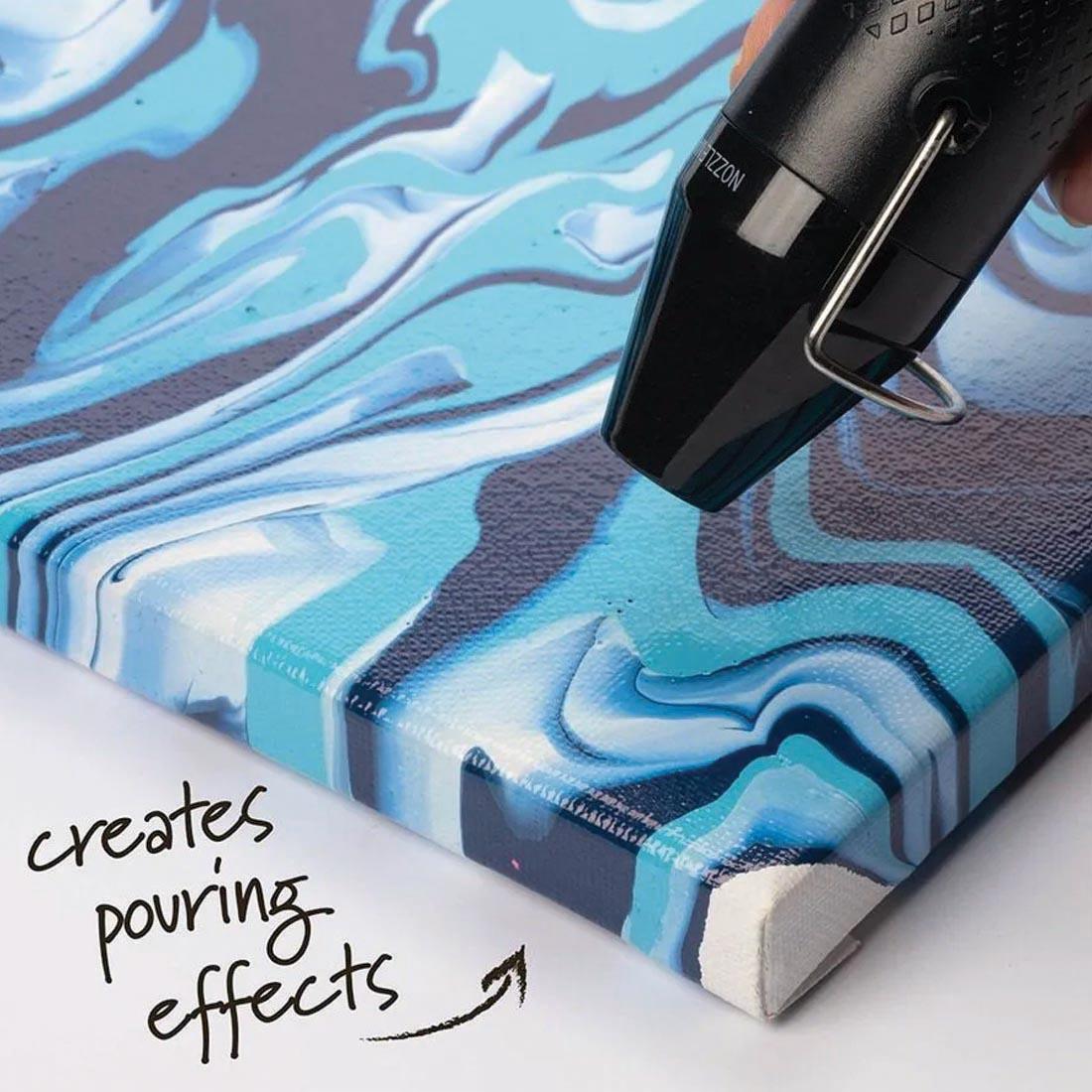 tip of Plaid Multi-Use Heat Pen hovering over a painted canvas with text: creates pouring effects