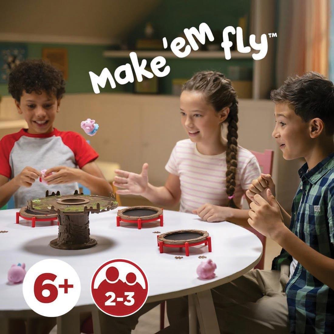 3 kids playing Pigs on Trampolines Board Game plus text make 'em fly; 6+; and symbol signifying 2-3 players