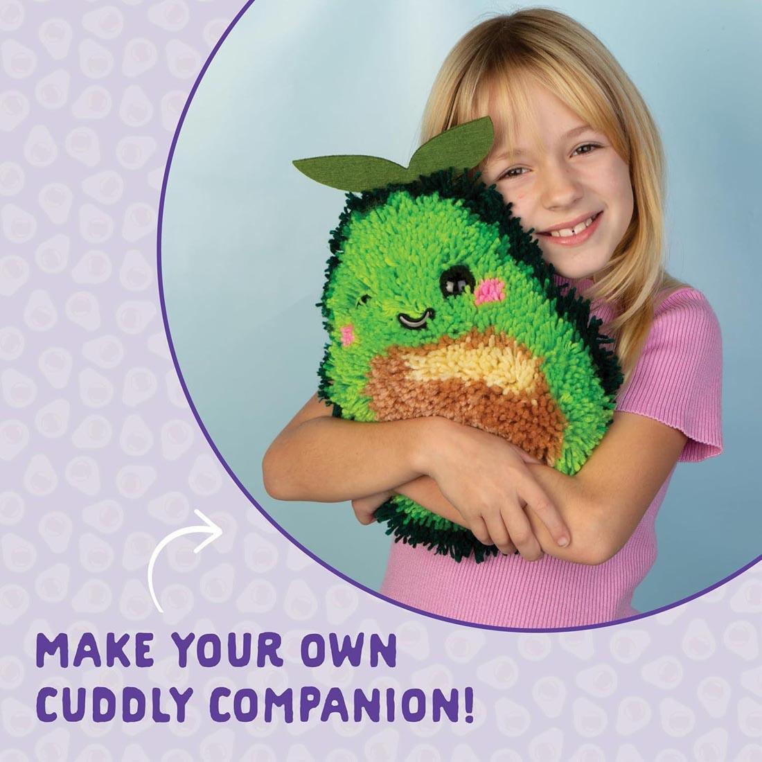 child hugging a completed Make An Avocado Latch Hook Pillow by Craft-Tastic plus text Make your own cuddly companion!