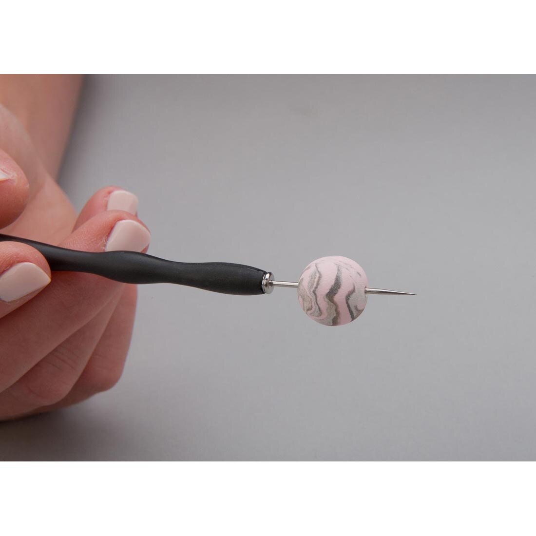 Hand holding a plastic tool with a needle end, with a round, marbled clay bead on the needle