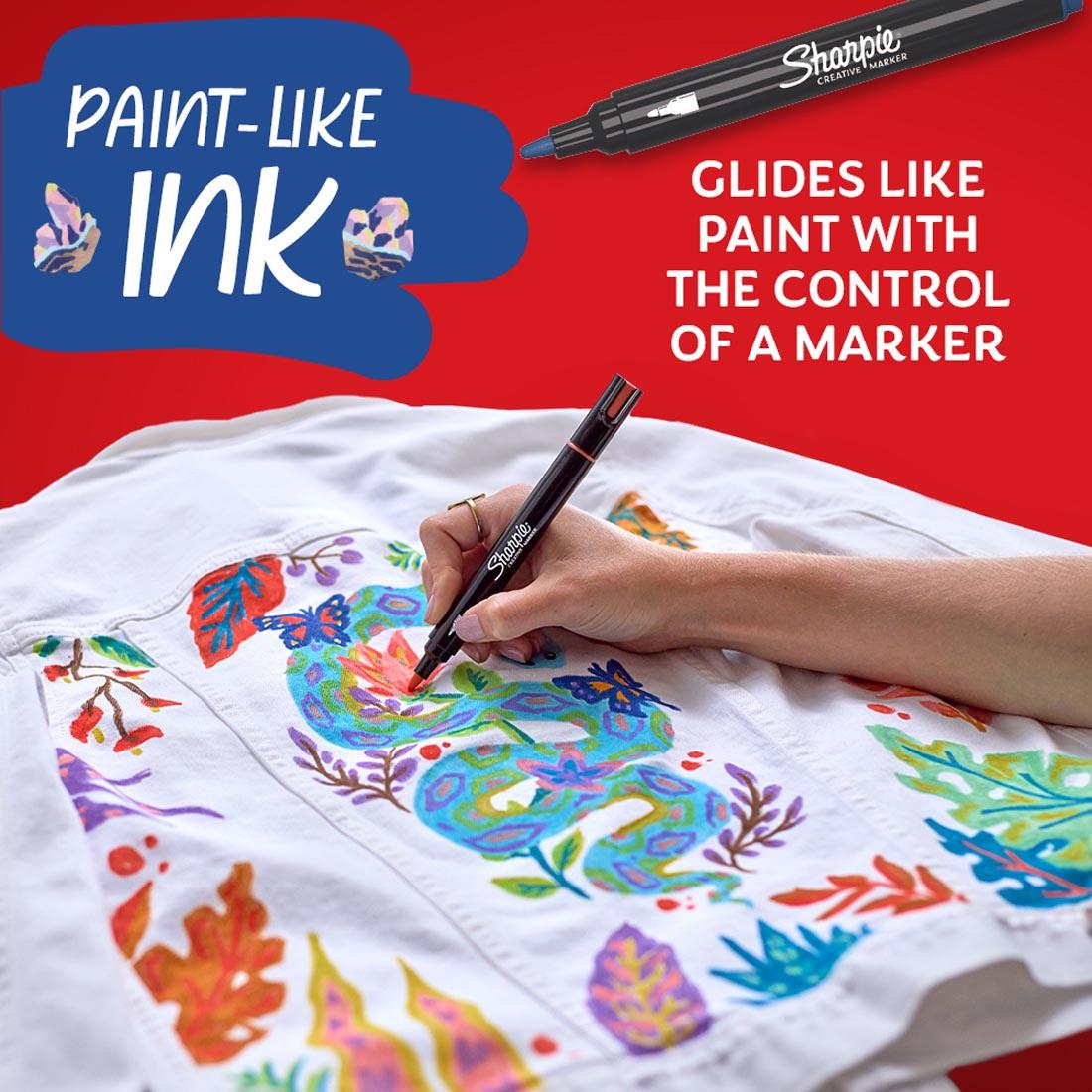 hand using a Sharpie Creative Bullet Tip Marker on white fabric plus text Paint-Like Ink; Glides like paint with the control of a marker