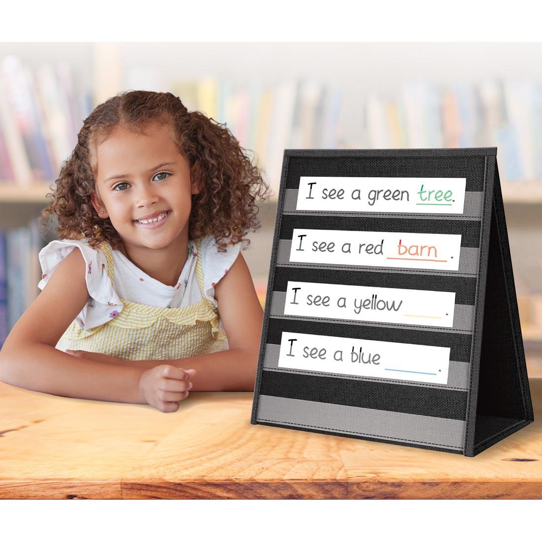 child sitting next to a Black Desktop Pocket Chart By Teacher Created Resources with completed sentence cards
