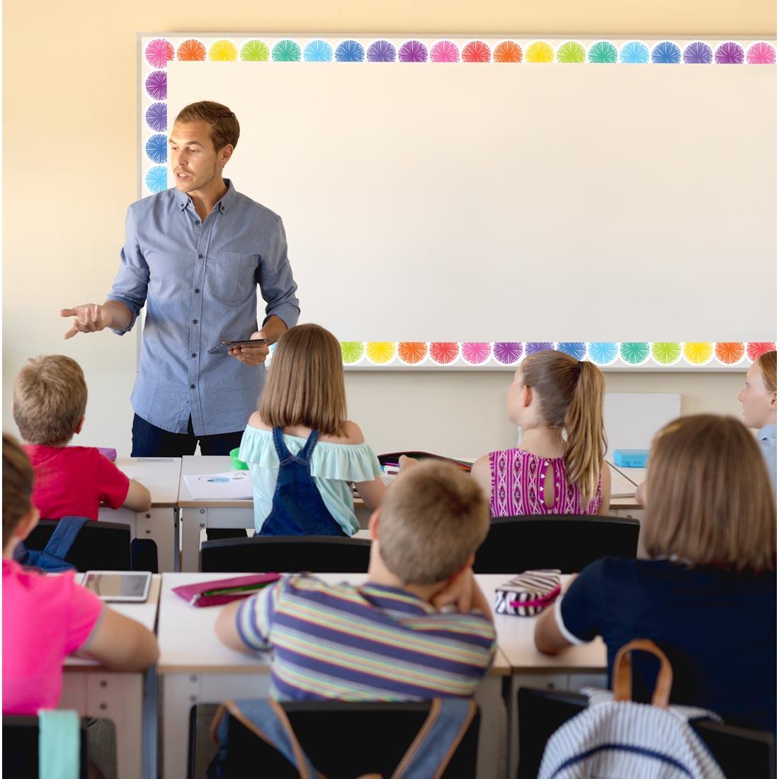 adult in a classroom in front of a board decorated with Poms Scalloped Die-Cut Border Trim from the Colorful collection by Teacher Created Resources