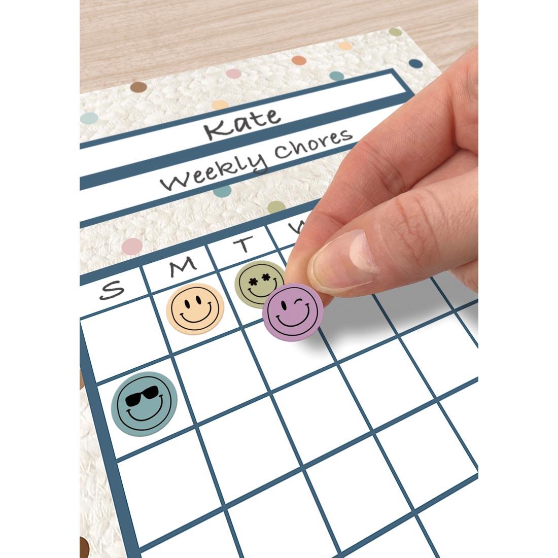 hand placing a Calming Colors Smiley Face Mini Sticker from the Everyone is Welcome collection by Teacher Created Resources on an incentive chart